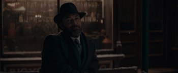 Movie still from “Fantastic Beasts: The Secrets of Dumbledore” (2022), directed by David Yates – A man wearing a suit and a hat sitting in front of a window; Medium shot, Over the shoulder angle