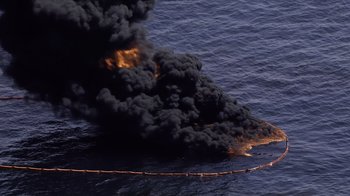 Movie still from “Fantastic Fungi” (2019), directed by Louie Schwartzberg – An oil spill in the middle of the ocean with black smoke billowing from it; Extreme Wide shot, Overhead angle