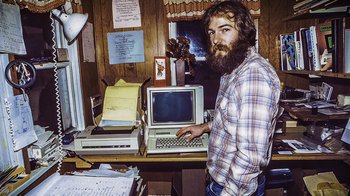 Movie still from “Fantastic Fungi” (2019), directed by Louie Schwartzberg – A man standing in front of a laptop computer on top of a desk; Medium shot, High angle