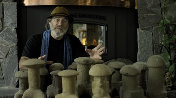 Movie still from “Fantastic Fungi” (2019), directed by Louie Schwartzberg – An older man is standing in front of a group of statues; Close Up shot, Overhead angle
