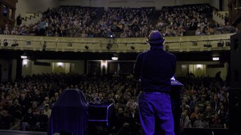 Movie still from “Fantastic Fungi” (2019), directed by Louie Schwartzberg – A man standing on a stage in front of an audience; Wide shot, High angle