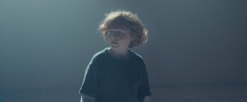 Movie still from “Firestarter” (2022), directed by Keith Thomas – A boy with red hair is standing in the dark; Close Up shot, Over the shoulder angle
