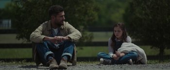 Movie still from “Firestarter” (2022), directed by Keith Thomas – A man sitting next to a little girl on the ground; Medium shot, Over the shoulder angle