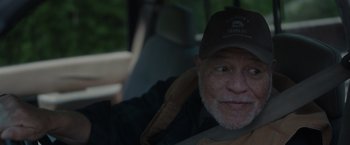 Movie still from “Firestarter” (2022), directed by Keith Thomas – An older man wearing a baseball cap sitting in the back of a car; Close Up shot, Over the shoulder angle