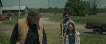 Movie still from “Firestarter” (2022), directed by Keith Thomas – A group of people standing on top of a grass covered field; Medium shot, Over the shoulder angle