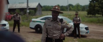 Movie still from “Firestarter” (2022), directed by Keith Thomas – A man in a uniform standing next to a white car; Medium shot, Over the shoulder angle