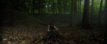 Movie still from “Firestarter” (2022), directed by Keith Thomas – A woman crouching in the woods near a pile of wood; Extreme Wide shot, High angle