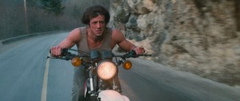 Movie still from “First Blood” (1982), directed by Ted Kotcheff – A man riding a motorcycle down the middle of the road; Medium shot, Low angle