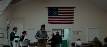 Movie still from “First Reformed” (2017), directed by Paul Schrader – A man with a long beard standing in front of an american flag hanging on the wall; Wide shot, Low angle