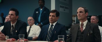 Movie still from “Flamin' Hot” (2023), directed by Eva Longoria – A man in a suit and tie sitting at a table with other men; Medium shot, Over the shoulder angle