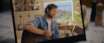 Movie still from “Flora and Son” (2023), directed by John Carney – A man sitting in front of a window playing a guitar; Medium shot, Low angle