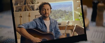 Movie still from “Flora and Son” (2023), directed by John Carney – A man sitting at a table holding a guitar; Close Up shot, High angle