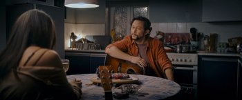 Movie still from “Flora and Son” (2023), directed by John Carney – A man sitting at a table with an acoustic guitar; Medium shot, Over the shoulder angle