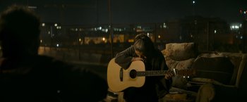 Movie still from “Flora and Son” (2023), directed by John Carney – A person holding a guitar while wearing headphones; Medium shot, Over the shoulder angle