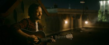 Movie still from “Flora and Son” (2023), directed by John Carney – A man holding a guitar in front of a building at night; Close Up shot, Over the shoulder angle