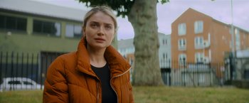 Movie still from “Flora and Son” (2023), directed by John Carney – A woman wearing a brown jacket standing next to a tree; Close Up shot, Low angle