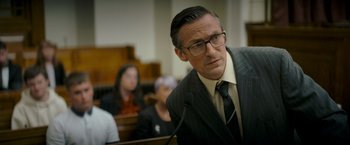 Movie still from “Flora and Son” (2023), directed by John Carney – A man in a suit and tie in front of a microphone in a courtroom; Medium shot, Low angle