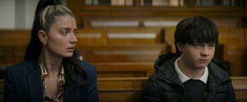Movie still from “Flora and Son” (2023), directed by John Carney – Two people sitting in a courtroom , one of whom is wearing a black jacket; Medium shot, Over the shoulder angle