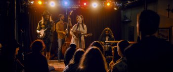 Movie still from “Flora and Son” (2023), directed by John Carney – A group of people sitting in front of a microphone on a stage; Wide shot, Low angle