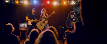 Movie still from “Flora and Son” (2023), directed by John Carney – A group of people sitting in front of a microphone and a guitar; Wide shot, Over the shoulder angle