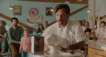 Movie still from “Flora & Ulysses” (2021), directed by Lena Khan – A man wearing a chef's hat and a white apron is preparing a doughnut; Medium shot, Over the shoulder angle