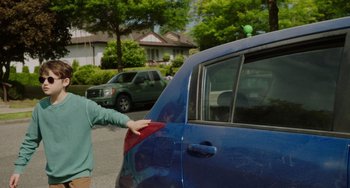 Movie still from “Flora & Ulysses” (2021), directed by Lena Khan – A person is putting a red seat belt on a blue car; Medium shot, Over the shoulder angle