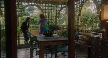 Movie still from “Flora & Ulysses” (2021), directed by Lena Khan – A woman sitting at a wooden table in the middle of a patio; Wide shot, Over the shoulder angle