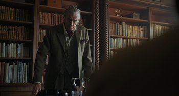 Movie still from “Flux Gourmet” (2022), directed by Peter Strickland – An older man standing in front of a bookshelf; Medium shot, Over the shoulder angle