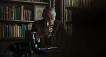 Movie still from “Flux Gourmet” (2022), directed by Peter Strickland – An older man sitting at a table with a glass of wine; Medium shot, Over the shoulder angle