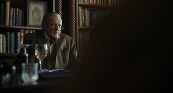 Movie still from “Flux Gourmet” (2022), directed by Peter Strickland – An older man sitting at a table with a glass of wine; Medium shot, Over the shoulder angle