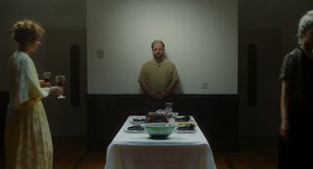 Movie still from “Flux Gourmet” (2022), directed by Peter Strickland – A man standing in front of a table with a bowl of cake; Medium shot, Over the shoulder angle