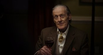 Movie still from “Flux Gourmet” (2022), directed by Peter Strickland – An older man holding a glass of wine in his right hand; Close Up shot, Low angle