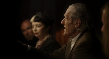 Movie still from “Flux Gourmet” (2022), directed by Peter Strickland – An older man and woman sitting next to each other in front of another man; Close Up shot, Over the shoulder angle