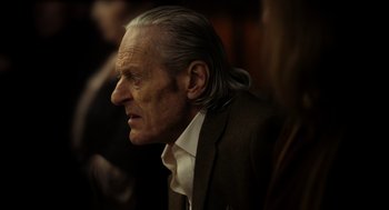 Movie still from “Flux Gourmet” (2022), directed by Peter Strickland – An old man with a long gray hair and wearing a suit; Close Up shot, Over the shoulder angle