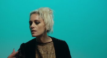 Movie still from “Flux Gourmet” (2022), directed by Peter Strickland – A woman with blonde hair is standing in front of a blue background; Close Up shot, Low angle