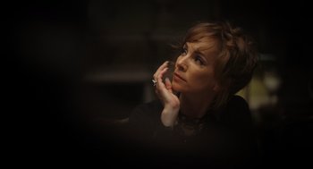 Movie still from “Flux Gourmet” (2022), directed by Peter Strickland – A woman sitting in a chair with her hand to her face; Close Up shot, Over the shoulder angle