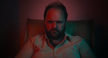 Movie still from “Flux Gourmet” (2022), directed by Peter Strickland – A bearded man sitting on a couch in a living room; Close Up shot, Low angle