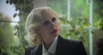Movie still from “Flux Gourmet” (2022), directed by Peter Strickland – A woman with blonde hair wearing a black suit and a white tie; Close Up shot, Over the shoulder angle