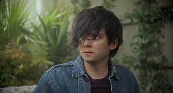 Movie still from “Flux Gourmet” (2022), directed by Peter Strickland – A young man with long black hair wearing a jean jacket; Close Up shot, High angle