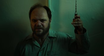 Movie still from “Flux Gourmet” (2022), directed by Peter Strickland – A man with a beard holding onto a chain; Close Up shot, Low angle