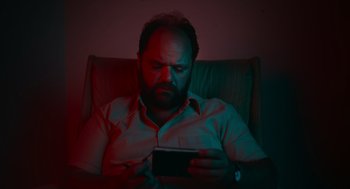 Movie still from “Flux Gourmet” (2022), directed by Peter Strickland – A man sitting on a couch looking at a phone; Close Up shot, High angle