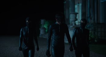 Movie still from “Flux Gourmet” (2022), directed by Peter Strickland – A group of people in black suits walking on a path at night; Medium shot, Low angle