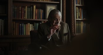 Movie still from “Flux Gourmet” (2022), directed by Peter Strickland – An older man sitting at a desk in front of a bookshelf; Medium shot, Over the shoulder angle
