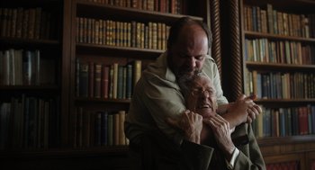 Movie still from “Flux Gourmet” (2022), directed by Peter Strickland – An older man and a younger man hugging in front of a book shelf; Medium shot, High angle