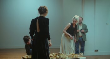 Movie still from “Flux Gourmet” (2022), directed by Peter Strickland – A group of people standing around a pile of bread; Medium shot, Over the shoulder angle