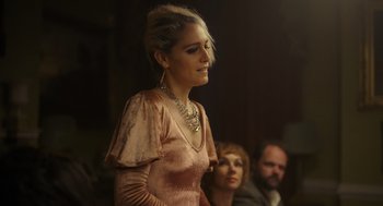 Movie still from “Flux Gourmet” (2022), directed by Peter Strickland – A woman in a pink dress standing in front of other people; Medium shot, Low angle