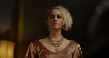 Movie still from “Flux Gourmet” (2022), directed by Peter Strickland – A woman wearing a necklace and earrings in front of a dark background; Close Up shot, Low angle