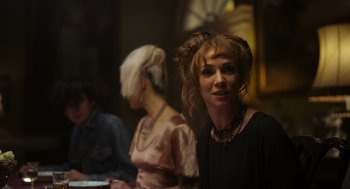 Movie still from “Flux Gourmet” (2022), directed by Peter Strickland – A woman in a black dress is sitting in front of other people; Close Up shot, Over the shoulder angle