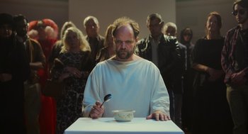 Movie still from “Flux Gourmet” (2022), directed by Peter Strickland – A man in white shirt holding a spoon in front of a bowl; Medium shot, Low angle