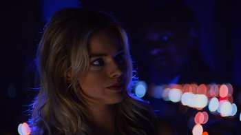 Movie still from “Focus” (2015), directed by John Requa – A beautiful blond woman with blue eyes and long blonde hair in front of blurry lights; Close Up shot, Over the shoulder angle